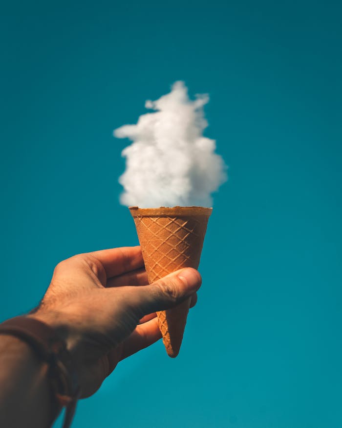 pexels-photo-1262302 Hand holding an ice cream cone with a cloud effect against a clear sky background.